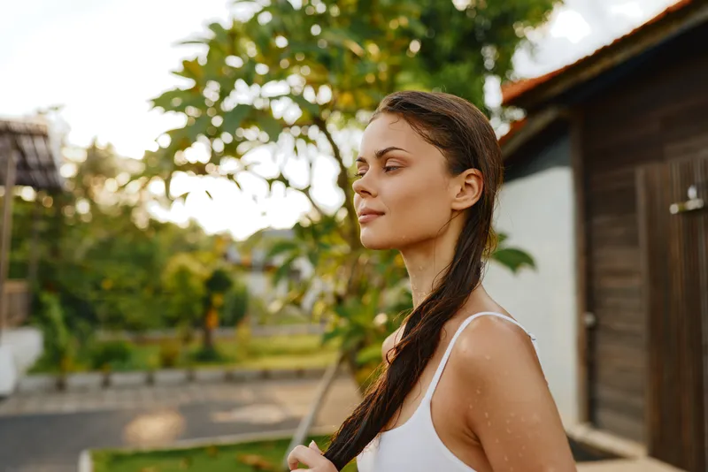 Young woman with long dark hair wearing a white tank top stands outdoors in a sunlit garden, looking to the side with a calm, reflective expression.