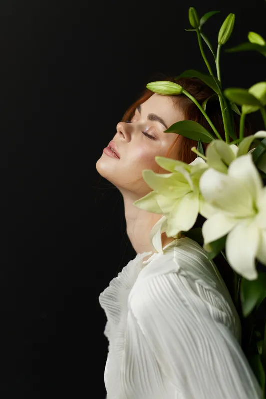 Elegant woman with red hair and floral arrangement, closed eyes exuding calmness, wearing a white blouse on a dark background that enhances the serene and sophisticated mood