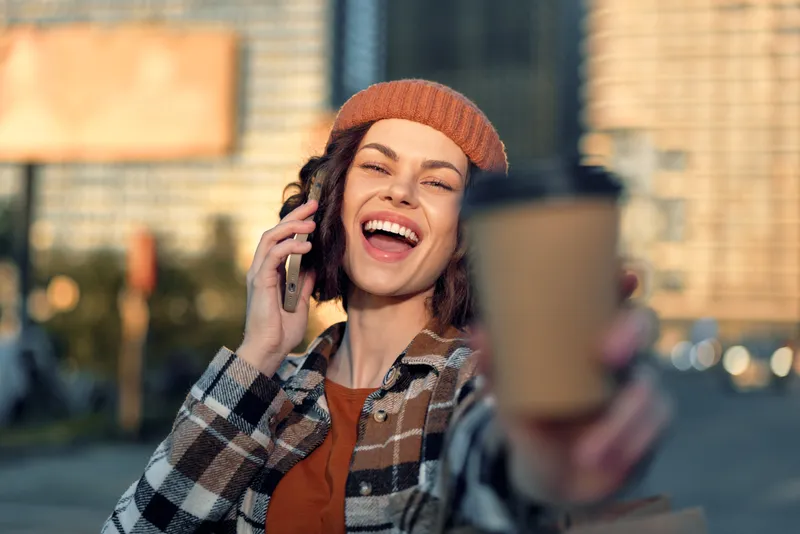 Woman holding coffee and phone with bright smile in urban street, candid lifestyle moment with beanie; golden hour glow, authenticity, mindful living and emotional storytelling vibe.