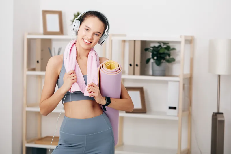 Fitness woman in sporty attire with headphones, holding a yoga mat and towel in a bright home setting, ready for a workout session and wellness routine.