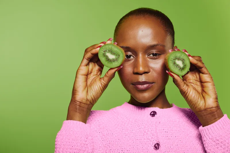 Portrait of a confident dark skinned African woman holding two halves of a kiwi fruit near her face. Bright green background and pink sweater create fresh, healthy lifestyle concept.