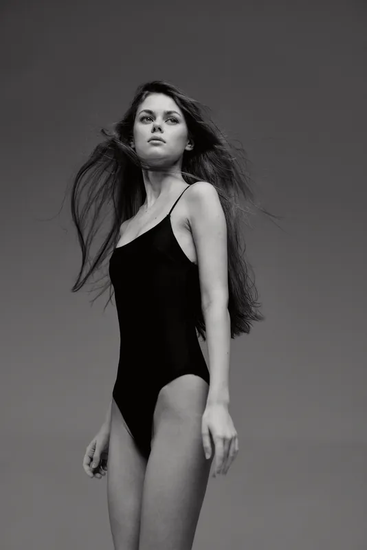 Black and white portrait of a young woman in a black swimsuit with flowing hair against a plain background