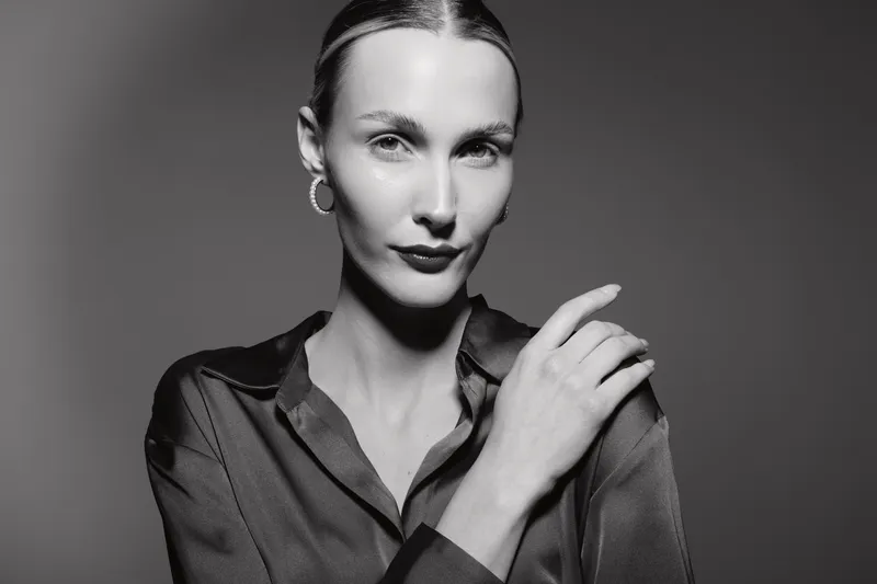 Portrait of a white woman with light skin and slicked hair wearing hoop earrings and dark shirt in studio, confident expression with hand near face.
