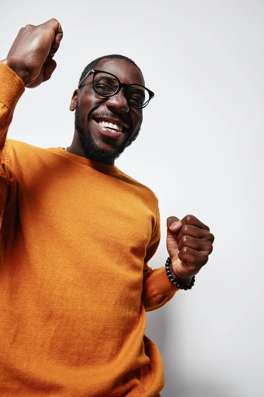 African man with dark skin wearing glasses and orange sweater smiling and celebrating with raised fists on white background. Happy male portrait with positive emotion.