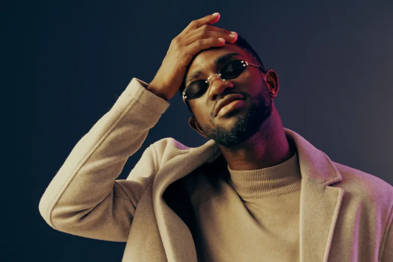 Stylish African man wearing sunglasses and a beige coat poses confidently against a colorful gradient background, showcasing modern fashion and self expression