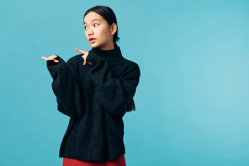 Young woman in black sweater with long dark hair pointing sideways and looking away on blue background. Female model wearing casual winter clothing with relaxed expression in studio.