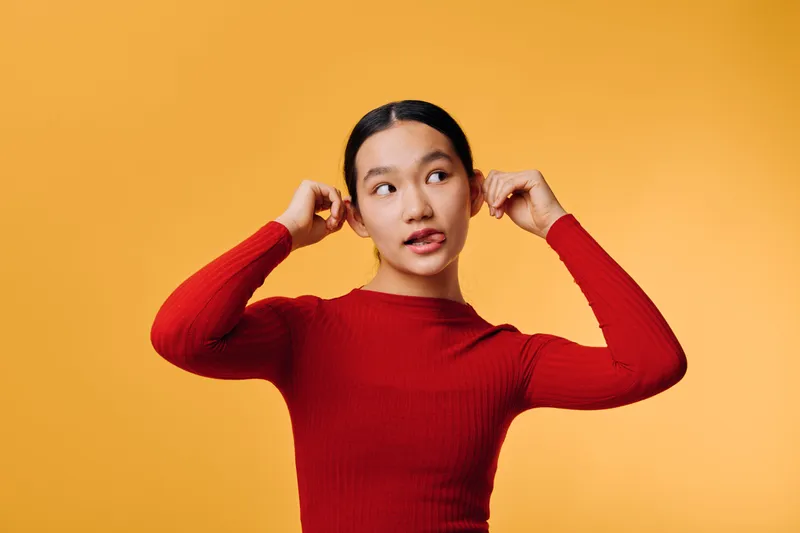 Young woman in red sweater posing against orange background stretching ears with fingers and looking to side with thoughtful expression, casual style and bright studio light.