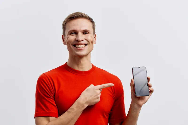 Smiling man wearing red t-shirt holding smartphone and pointing at it isolated on plain light gray background studio portrait. People lifestyle concept