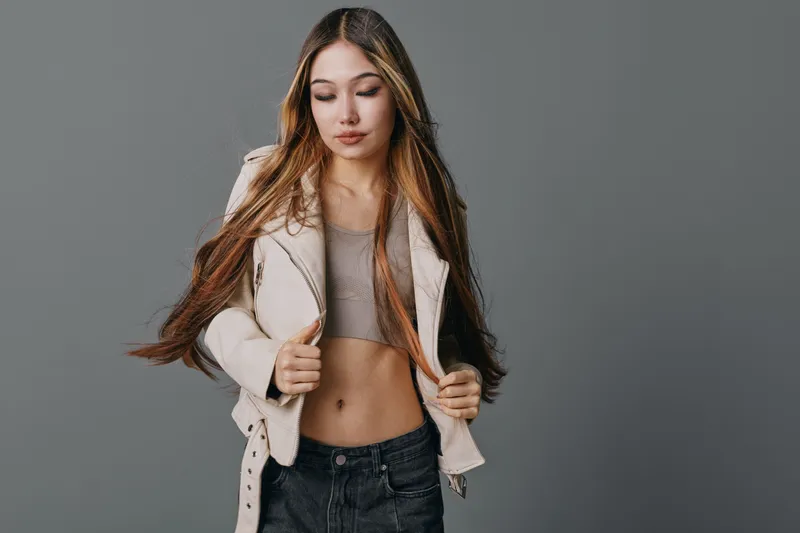 Young woman with long brown hair wearing a cropped top and light jacket posing in studio. Casual fashion outfit with confident expression and natural makeup on gray background.