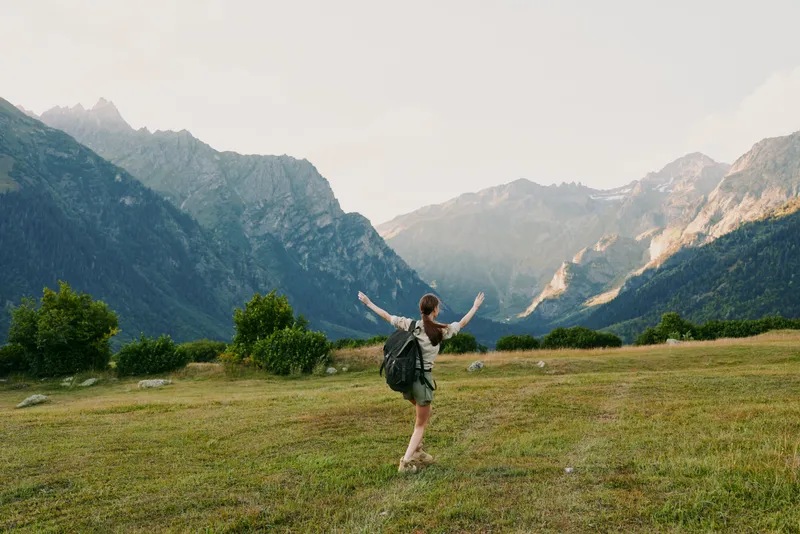 A young woman with a backpack joyfully walks across a grassy field surrounded by mountains under a bright sky, enjoying an outdoor hiking adventure.