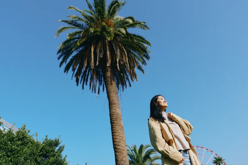 Woman beside a tall palm tree under a clear blue sky, wearing a light jacket and flowing dress, enjoying a sunny outdoor fashion moment and breeze