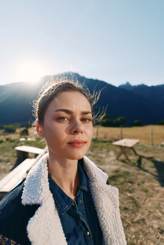 Woman portrait outdoors with mountains and sunlight in background, closeup face and natural expression in a field, wearing warm jacket and fleece collar for a scenic hike.
