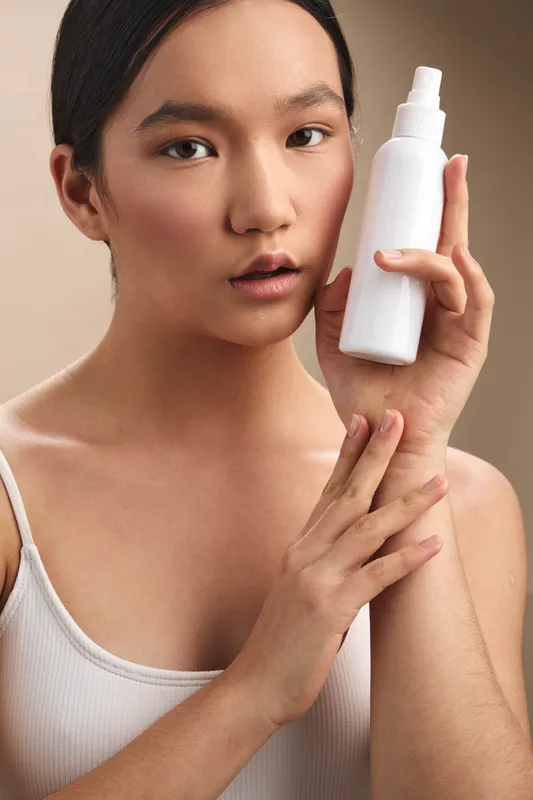Asian woman applies skincare product in a clean, bright setting. She holds a white bottle near her face, looks calm and focused, creating a serene and reassuring skincare moment.