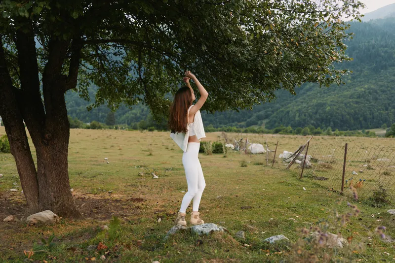 Young woman in white outfit standing under a tree in a rural field, natural light, relaxed pose, green landscape, casual lifestyle moment
