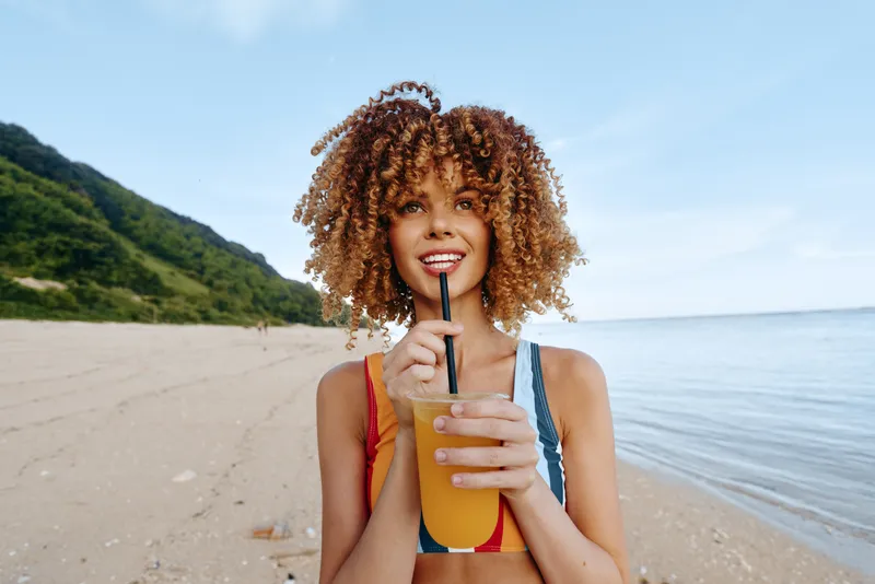 Woman at beach, ocean summer breeze, curly hair smiling in bikini, holding drink straw bottle near shoreline, sunny sky, coastal mountains, relaxed vacation mood, natural colors outdoors