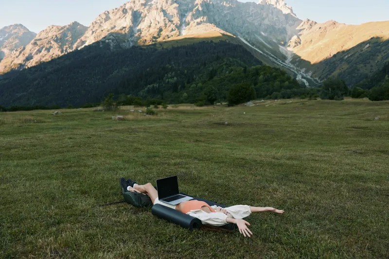 A person rests on a grassy hillside with a laptop, enjoying a peaceful outdoor workspace. Mountain scenery surrounds them, creating a calm, productive atmosphere in nature.