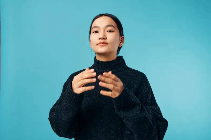 Young woman with dark hair wearing black sweater posing with hands in front of neutral blue background studio portrait of confident female with natural makeup and casual style.