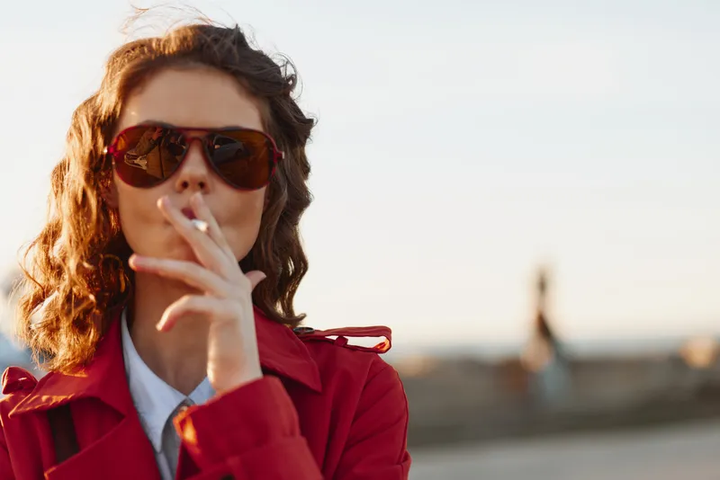 A stylish young woman in a red coat and sunglasses smokes a cigarette on an outdoor boardwalk by the beach, posing for a candid contemporary street scene.