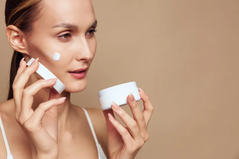 A young woman applies white cream on her cheek from a jar while holding a tube in her other hand. The neutral background highlights skincare and beauty care concepts.