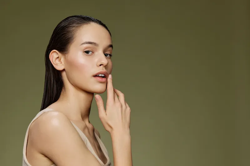 Elegant young woman with natural makeup touching her lip, set against a soft green background, emphasizing beauty, confidence, and self care
