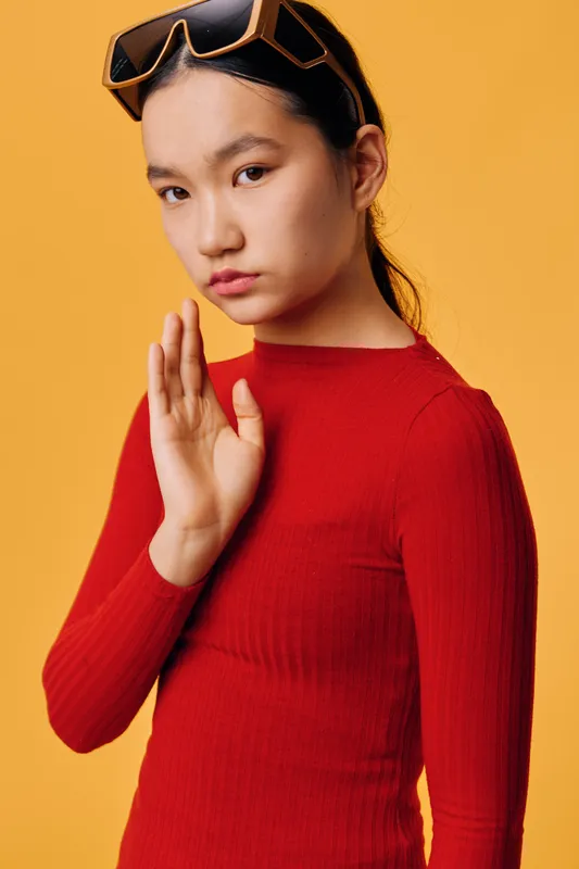 Young woman in red sweater with sunglasses on head, raising hand in stop gesture, looking serious against orange background in studio, expressing refusal or disagreement with direct eye contact.