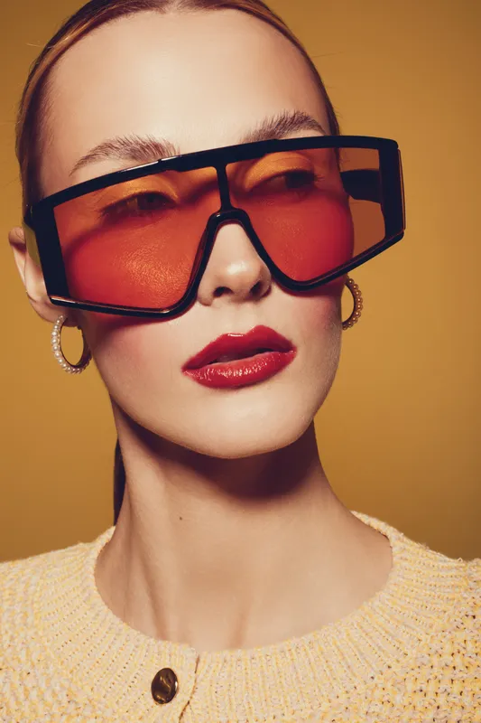 Close-up portrait of a stylish woman wearing oversized orange sunglasses and gold hoop earrings. She has glossy red lips and a beige knitted sweater on a warm brown background.