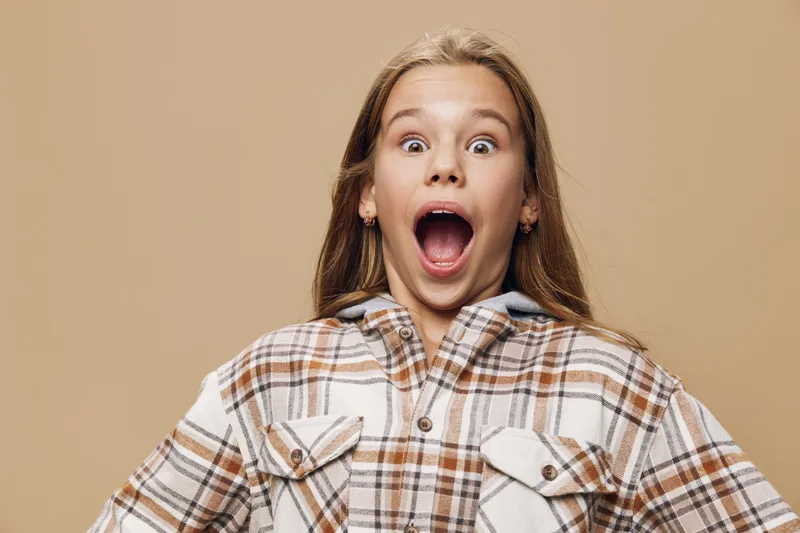 Girl with long hair wearing plaid shirt showing surprised expression with wide open mouth and raised eyebrows on beige background, casual clothing and emotion concept.