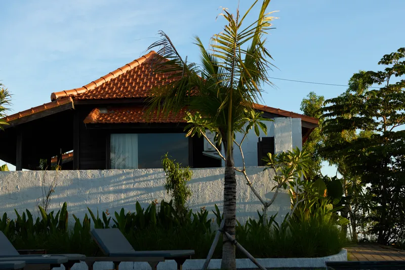 Tropical villa retreat scene with palm trees, white walls, and lounge chairs. Bright sunshine and calm ambiance promote relaxation, leisure, and a luxurious holiday mood.