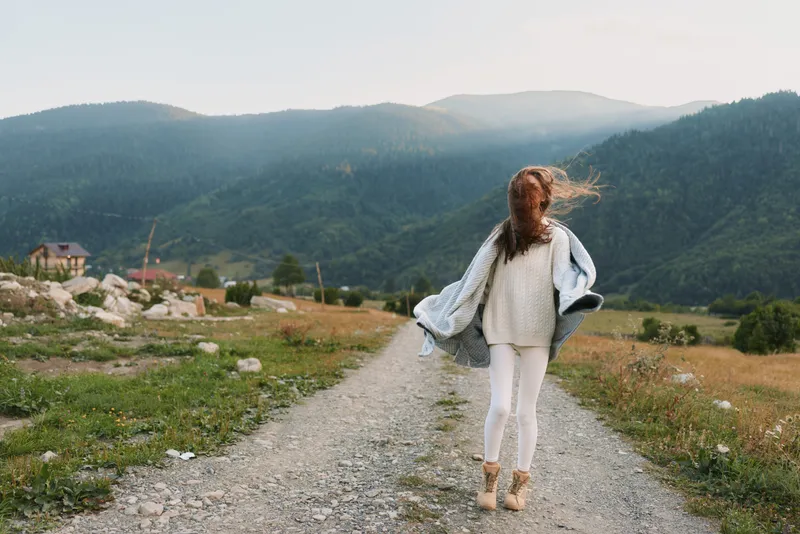 Casual woman walking along a rural gravel path, countryside landscape with distant mountains, soft breeze, light sweater and jeans, relaxed mood, sunlit atmosphere, natural colors.