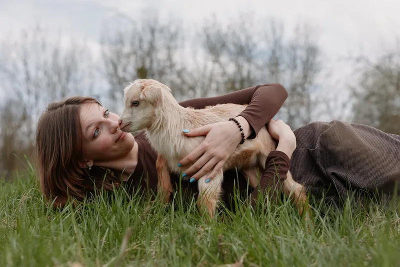A relaxed woman lies on grass, gently cradling a young goat as they share a tender moment outdoors. A lifestyle image depicting companionship, nature, and the bond between human and animal.
