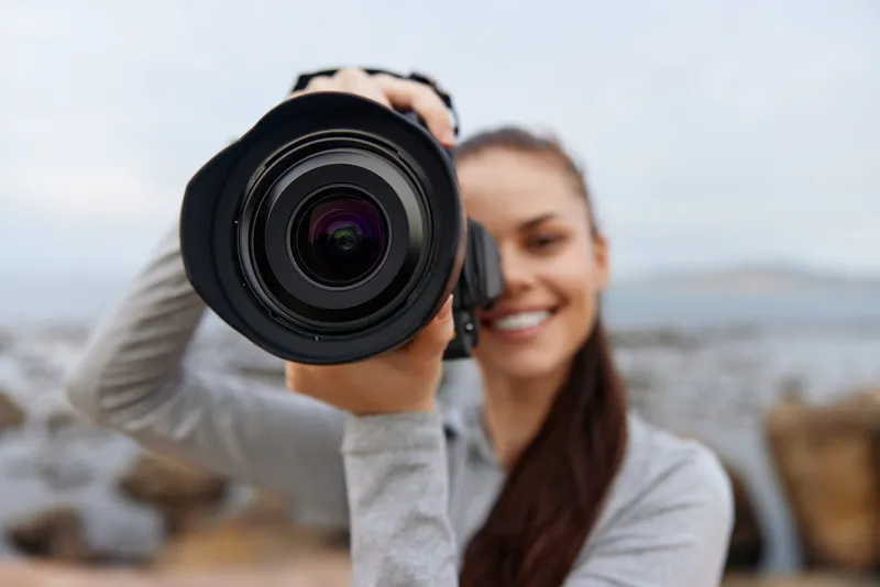 Young woman smiling while holding a camera outdoors, showcasing photography passion against a scenic backdrop, perfect for creative projects and social media
