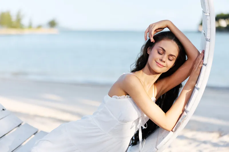 A young woman in a white dress rests against a curved seaside bench, eyes closed, hair flowing, basking in soft sunlight beside tranquil water.