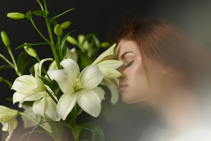 A serene young woman of Caucasian descent gently touching her face to white lilies, surrounded by a soft, ethereal atmosphere, representing tranquility and nature