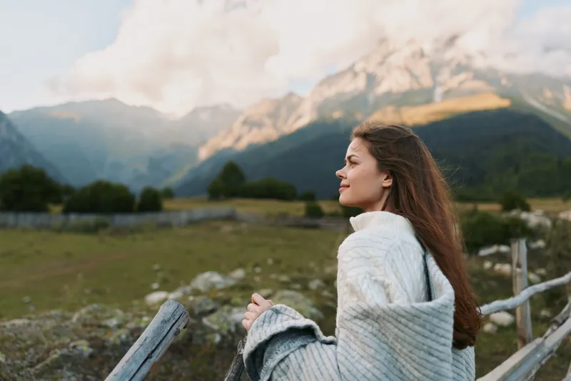 Young woman with long curly hair enjoys peaceful mountain landscape while wrapped in cozy knitted sweater standing near wooden fence in early autumn.