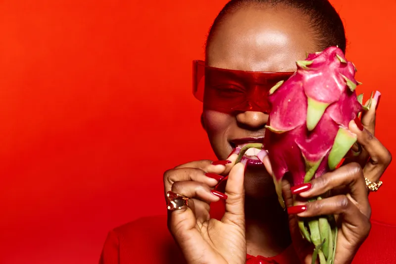 Dark skinned African woman in red outfit holds dragon fruit near face, smiling with closed eyes. Bright red background emphasizes vibrant mood and exotic fruit concept.