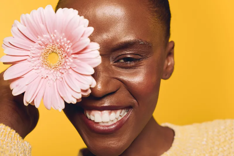 Smiling dark skinned African woman holds pink flower near face on yellow background. Concept of natural beauty, happiness, skincare, and floral wellness with bright studio lighting.