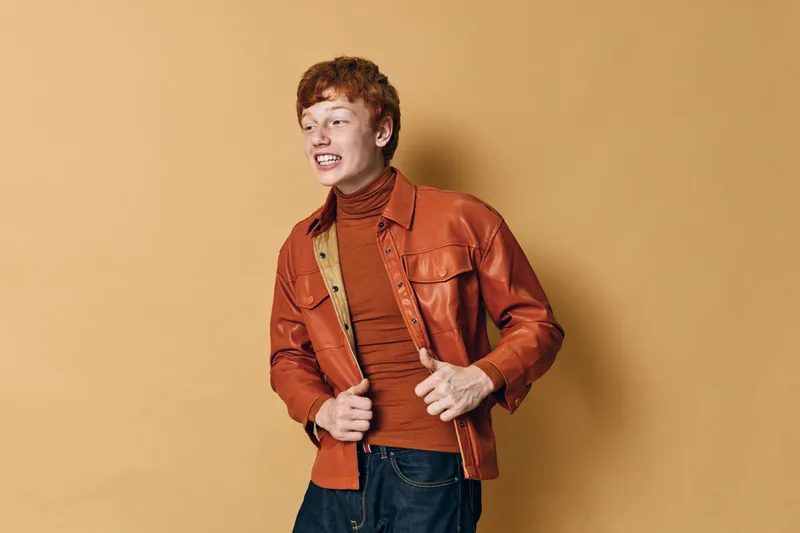 Young man with red hair wearing orange jacket and turtleneck smiling against beige background in casual style. Portrait of happy male model in studio with trendy outfit and expressive face.