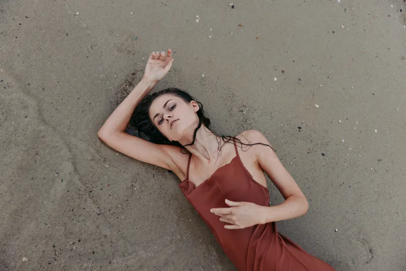 Sunset-lit portrait of a woman in a red slip dress lying on damp sand, eyes closed, calm expression, relaxed pose, gentle breeze, soft shadows, cinematic mood, intimate beach scene.