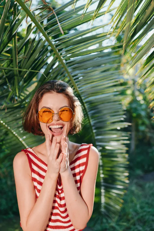 A cheerful woman poses outdoors in a tropical garden. She wears a striped top, round orange sunglasses and smiles brightly, creating a vibrant, carefree summer moment.