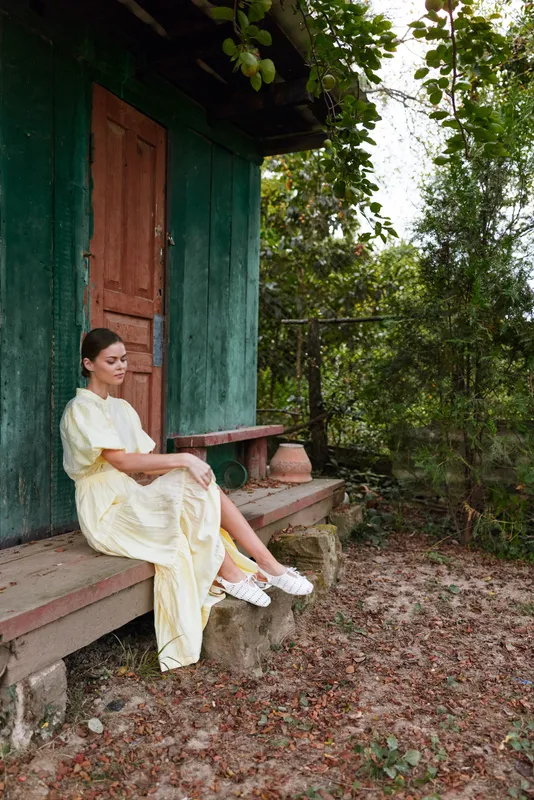 A woman sits on a rustic porch beside a green house, wearing a light yellow dress, creating a peaceful rural scene with natural surroundings and gentle afternoon light