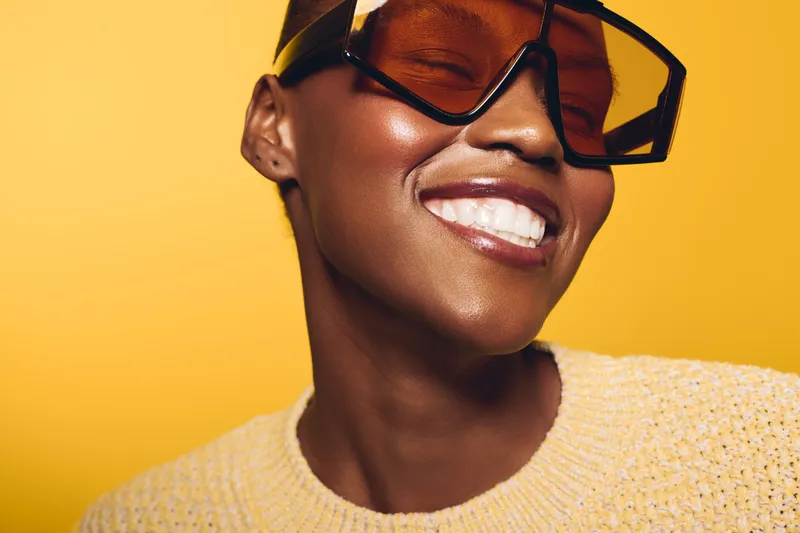 Close-up of smiling black African woman with dark skin wearing large sunglasses and yellow sweater on bright yellow background. Concept of happiness, fashion, and beauty portrait.