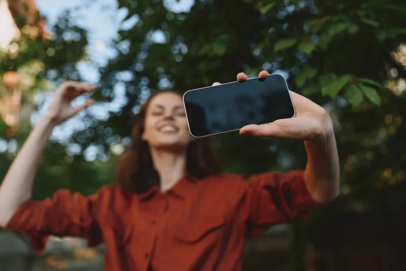 A young brunette woman takes a selfie with her phone outdoors, smiling as she extends her arm toward the camera in a casual everyday moment