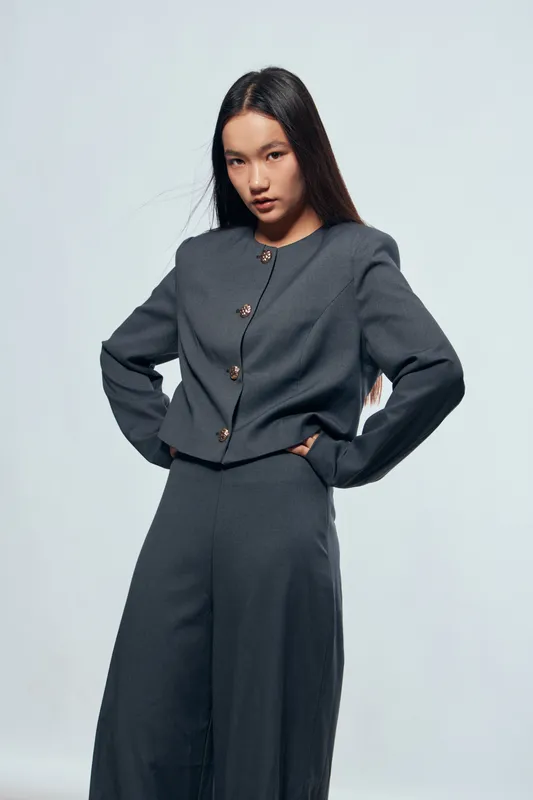 Young woman with long dark hair wearing gray pantsuit stands confidently with hands on hips against light background. Fashion model, portrait, elegance and style in clothing studio.