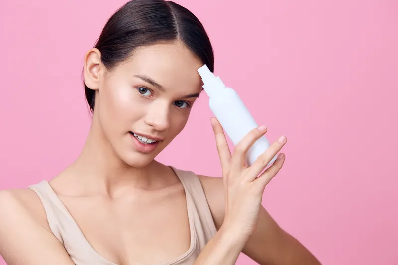 Young woman demonstrating a skincare product against a pink background, showcasing healthy skincare and beauty routines with an elegant expression