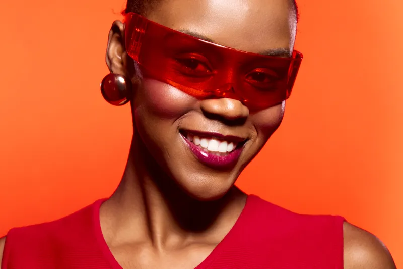 Close-up portrait of a smiling black woman wearing red sunglasses and red top against orange background. Concept of fashion, confidence, beauty, and vibrant style for dark skin models.