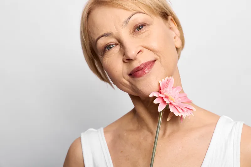 Woman portrait with a pink flower at her neck, blonde smile under studio light, soft skincare and natural beauty makeup, minimal background, fresh spring mood, close headshot