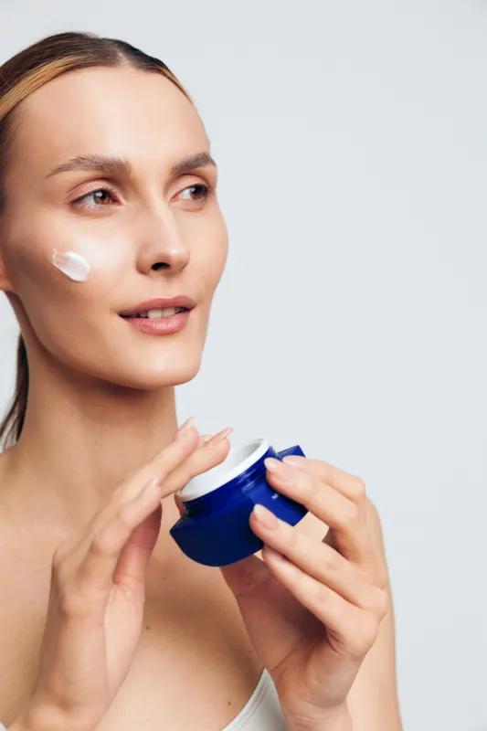 Young woman applies moisturizing cream on her face holds blue jar skincare beauty routine natural skin care isolated on white background with positive facial expression