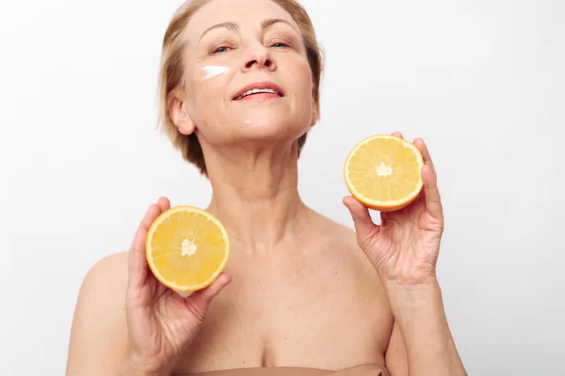 Mature woman with short hair holding two orange halves applies natural face cream on skin, smiling gently on white background. Concept of skincare, aging, and healthy lifestyle.