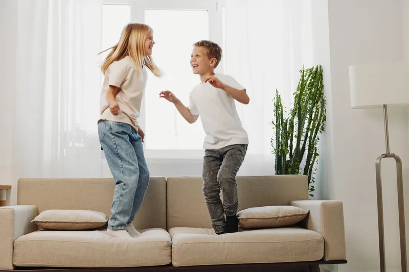 Two happy children jumping on beige sofa in bright living room with natural light and houseplant near window. Fun home activity and childhood joy concept.