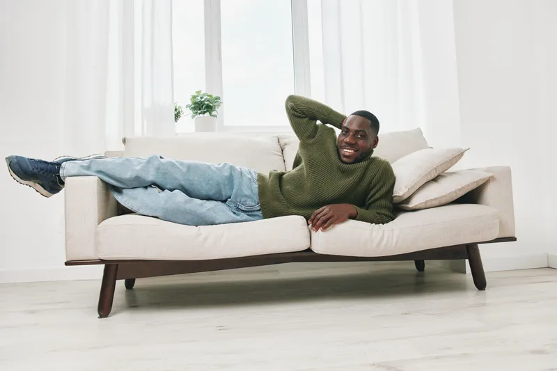 Smiling African American man lounging on a modern couch in a cozy, bright living room, showcasing relaxed ambiance and contemporary design